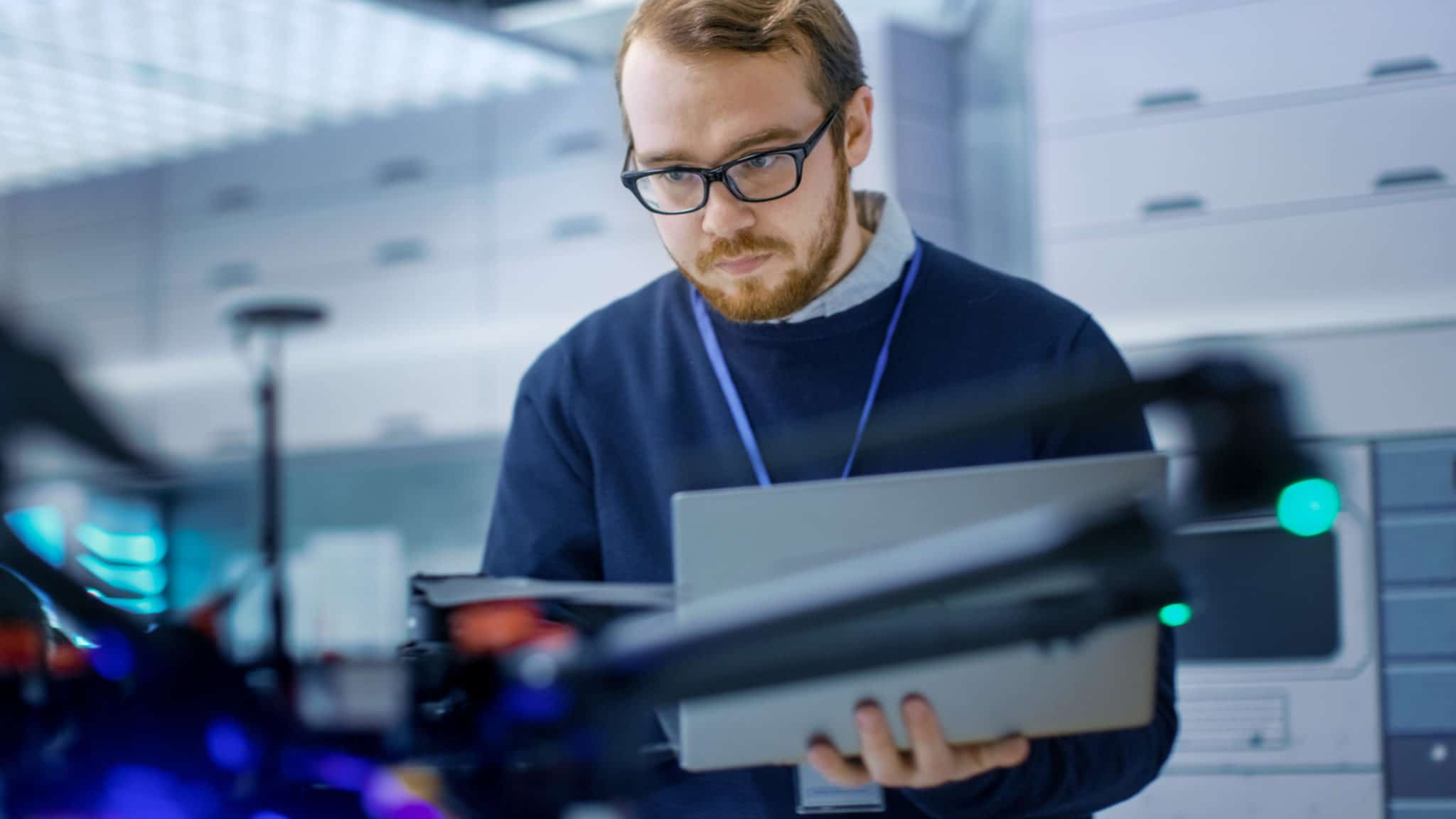 Engineer wearing glasses and blue sweater golding laptop computer while looking at drone.